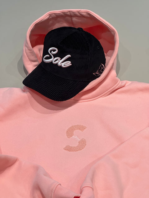 Sole Savaz black snapback cap placed inside the hood of the matching pastel pink towel-embroidery hoodie, showcasing the identical pink color tones.