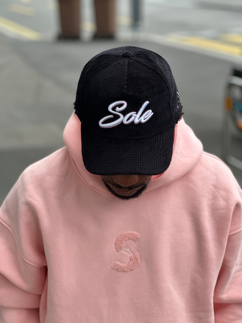 Front-facing lifestyle shot of the Sole Savaz black corduroy cap with white 3D "Sole" branding and pink accent stitching, paired with the signature pink hoodie.